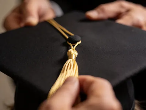 Close-up of a golden tassel tied to a black graduation cap with hands adjusting the knot against a blurred background