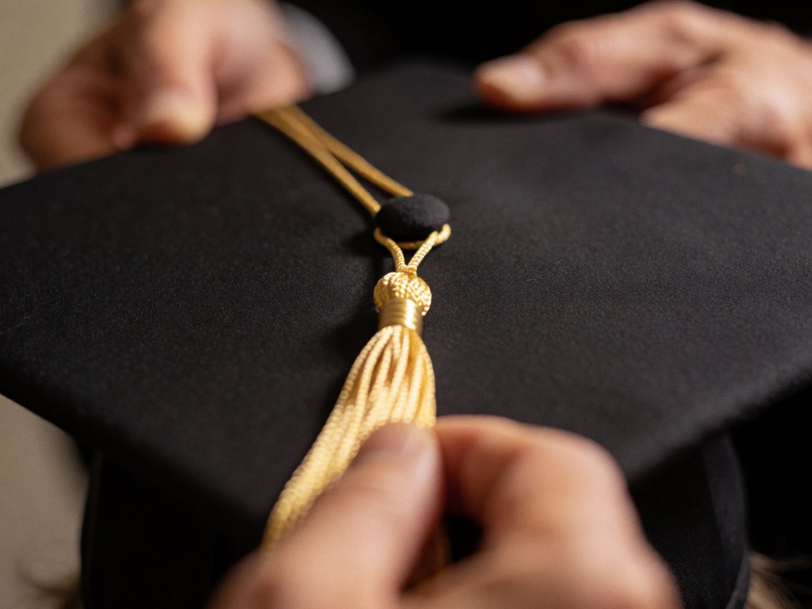 Close-up of a golden tassel tied to a black graduation cap with hands adjusting the knot against a blurred background