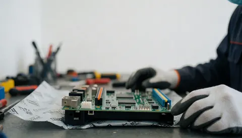Technician wearing gloves repairing a washing machine control board on a workbench in a workshop with blurred tools in the background