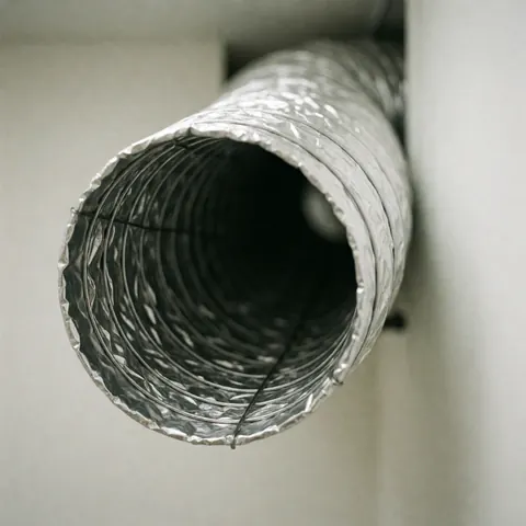 Close-up of a flexible HVAC air duct with exposed wire reinforcement, mounted on a white wall for heating repair work