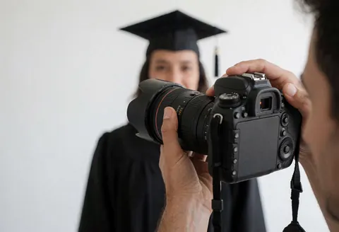 Professional photographer adjusting focus ring on DSLR camera to capture a graduate in academic gown against white background with soft natural light.
