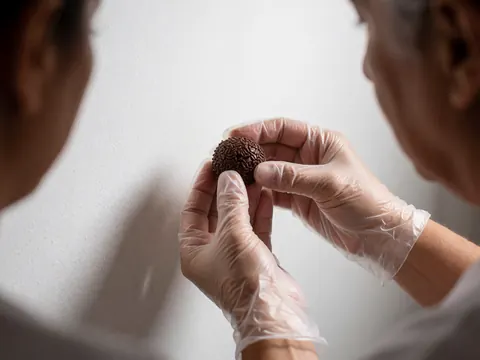 Gloved hands placing a chocolate brigadeiro covered in sprinkles on a white background, close-up detail of the dessert and granules.