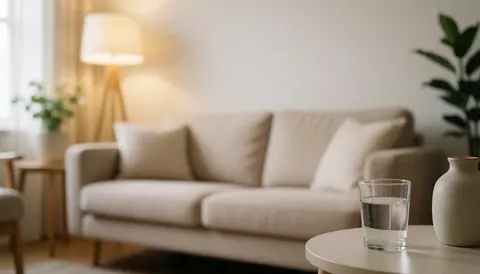 Warm blurred background of a living room with beige sofa, lamp, and table with water glass and vase, ideal for text overlay