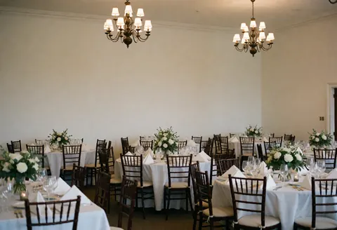 Empty wedding event space featuring white tablecloths, wooden chairs, floral centerpieces, and hanging chandeliers illuminating the elegant interior.