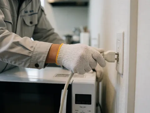 Technician wearing glove plugs microwave power cord into kitchen outlet with blurred background
