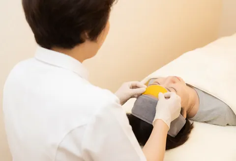 A therapist wearing gloves applies a yellow fruit slice to a client's forehead during a facial hydration treatment in a warm beige room.