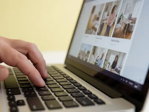 Close-up of an adult hand typing on a laptop keyboard with a photo gallery displayed on the screen against a pale yellow wall.