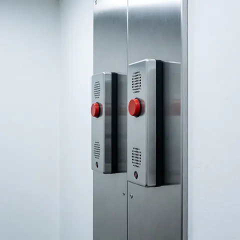 Two emergency intercoms with red buttons mounted on a metal panel in an elevator room. Clean cold lighting, white walls, and robust industrial design.