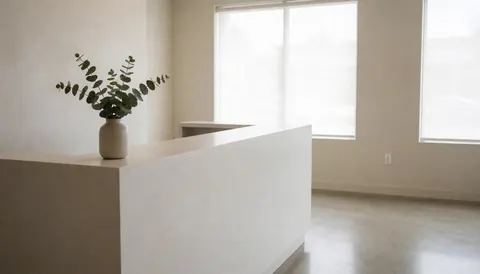 Minimalist dermabrasion clinic reception featuring a white desk, potted eucalyptus plant, and large windows with natural light creating a serene and clean atmosphere.