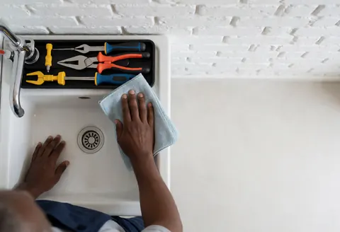Plumber cleaning white sink with microfiber towel, tools on tray, white brick wall, bright lighting, text space on right