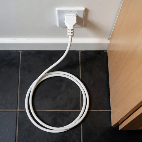 Close-up view of a white electrical cord plugged into a wall outlet in a kitchen with dark tiled floor and wooden cabinetry