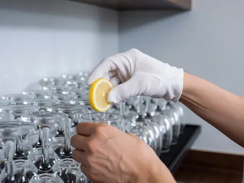 Close up of a white gloved hand placing a lemon slice into a clear bar glass with wine glasses in the background