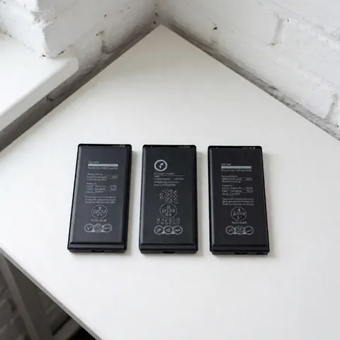 Three new sealed cell phone batteries with technical labels on a white table against a white brick wall with natural lighting
