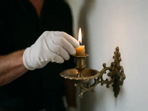 A gloved hand adjusts the flame of a beeswax candle on an antique bronze candelabra against a dark background and white wall