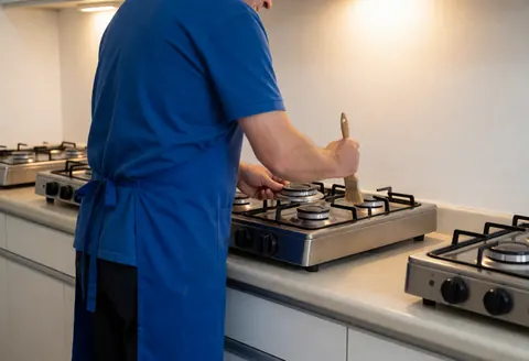 Technician in blue uniform cleaning a stove burner with a brush in a kitchen with white walls and warm lighting