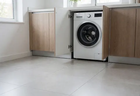 White front-loading washing machine integrated into a modern kitchen cabinet with gray tile flooring and natural light from a window.