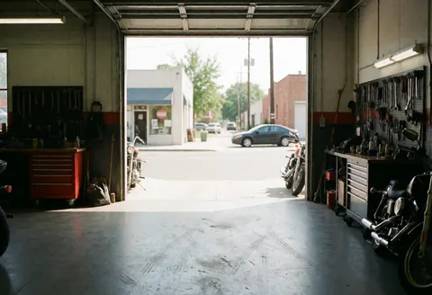 Motorcycle garage interior featuring tools mounted on the wall, concrete floor with tire marks, parked motorcycles, and a street view with cars in the background.