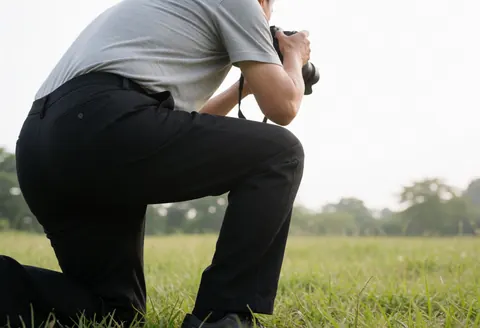 Professional wedding photographer kneeling in a green field, capturing images with a DSLR camera under soft natural light during an outdoor session.