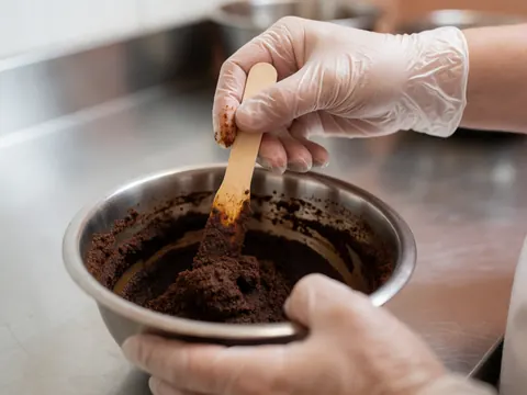 A gloved hand mixes coffee and cocoa paste in a metal bowl on a stainless steel table under clean clinical lighting.