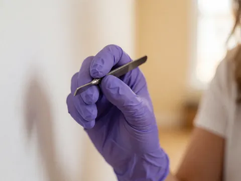 Close-up of a hand wearing a purple glove holding a precision eyebrow lamination tool against a warm, blurred indoor background.