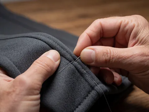 Close-up of an adult hand stitching gray fabric with a needle against a wooden background, highlighting visible texture and professional precision.