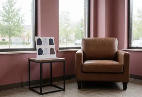 Brown leather chair and shirt catalog on table in waiting room with large windows and dusty rose walls