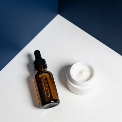 Amber glass serum bottle and white cream jar on white surface with blue walls, soft clean lighting for beauty products
