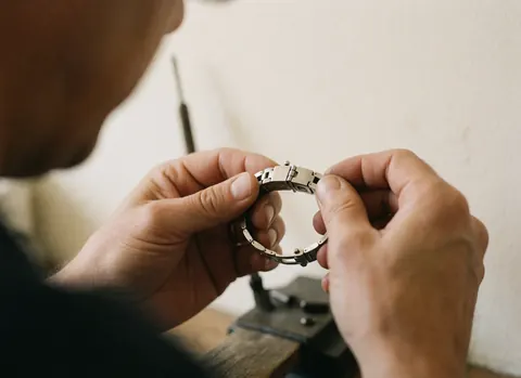 Close-up of hands repairing a metal watch clasp with precision tools in a bright workshop with blurred white walls