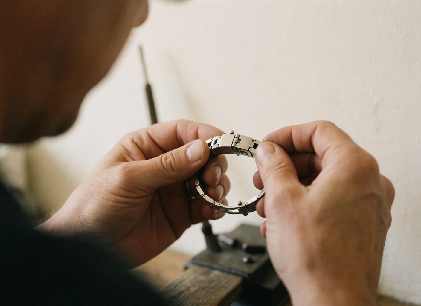 Close-up of hands repairing a metal watch clasp with precision tools in a bright workshop with blurred white walls