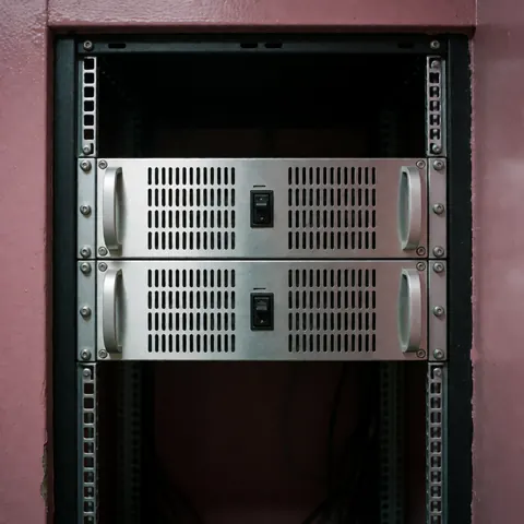 Two professional metal audio amplifiers with ventilation mounted in a 19-inch rack against a pink wall with cool technical lighting