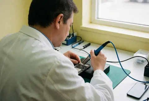 Close-up of a technician using a soldering iron to repair a mobile phone circuit board on a workbench with tools and natural lighting