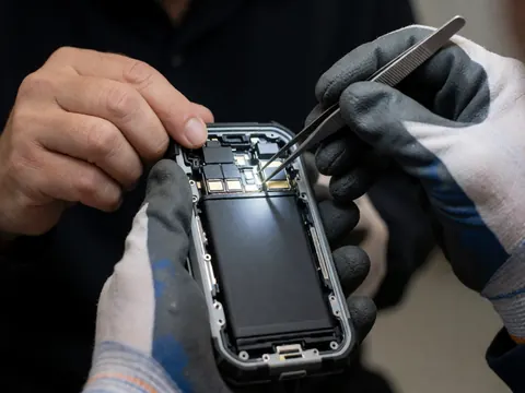 Technician wearing gloves cleans internal contacts of a rugged phone with tweezers, bright lighting on metal components against a dark background