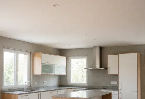 Open modern kitchen featuring white cabinets, granite countertops, concrete walls, and windows with natural light, ideal for kitchen appliance repair services.
