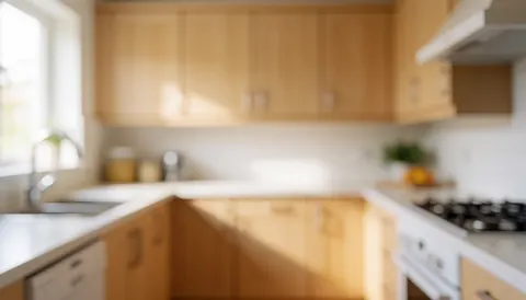 Residential kitchen featuring light wood cabinets, a white countertop, and appliances with a blurred background for text overlay and warm natural lighting.