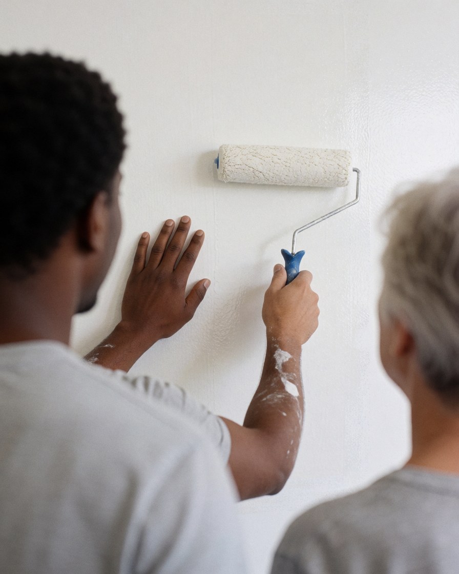 A painter uses a roller to apply fresh white paint to an interior wall while a client watches from behind, showing a clean and bright finish.