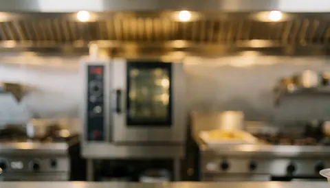 Out-of-focus commercial kitchen featuring stainless steel ovens and ventilation under warm lighting, suitable for text overlay