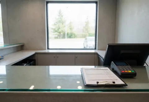 Reception counter in a mobile repair shop featuring a payment terminal, clipboard form, concrete walls, and a window with natural light.