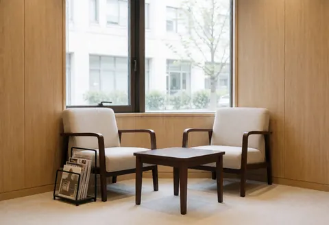 A waiting area featuring upholstered armchairs, a coffee table, and a magazine rack near a window with natural light.
