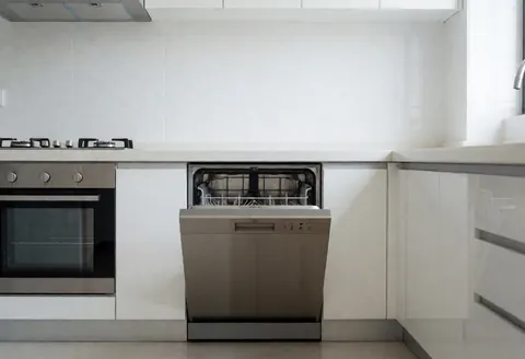 Open built-in dishwasher in a modern kitchen with empty racks, white tile walls, and stainless steel appliances