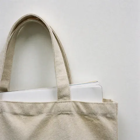 Beige canvas tote bag with visible stitching, white notebook peeking out, white background, even lighting