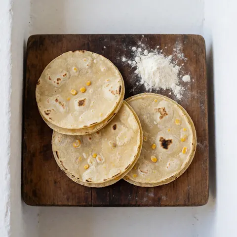 Freshly made corn tortillas with visible grains on a dark wooden board sprinkled with corn flour, lit by natural light.