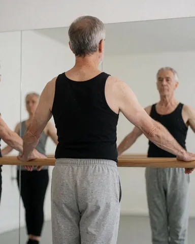 Back view of an older man holding a wooden ballet barre in a dance studio with a mirror reflecting his posture against a white wall.