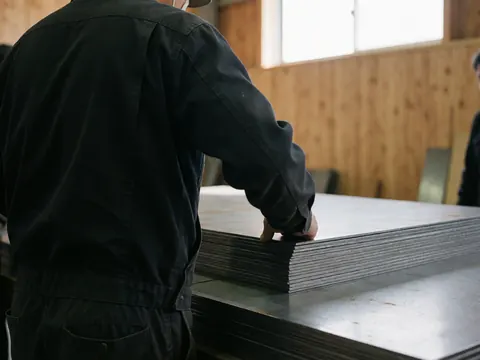 A worker carefully inspects a stack of cut steel plates in a metal workshop with natural light and wooden walls.