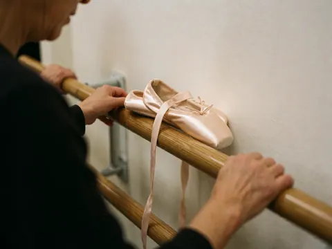 Close-up of pink ballet shoes resting on a polished wooden barre with hands adjusting the equipment in a bright studio