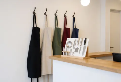 Dark and light colored kitchen aprons hanging on hooks against a white wall with wooden countertop and cooking tools nearby