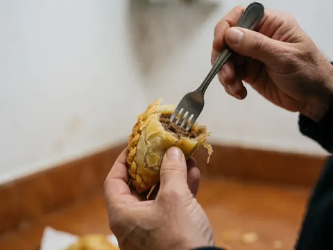 Close-up of a hand using a fork to break open a golden empanada, revealing juicy meat filling with a blurred kitchen background.