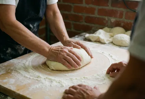 Professional hands kneading dough on a floured wooden board with a red brick wall background in a kitchen workshop setting