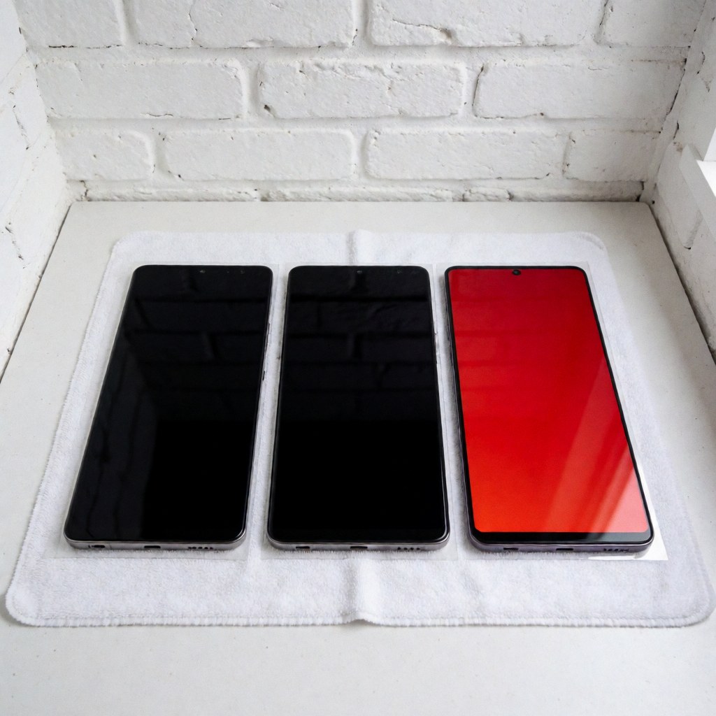 Three new smartphones on a white towel against a white brick wall with even lighting, one displaying a bright red screen, ideal for mobile repair services