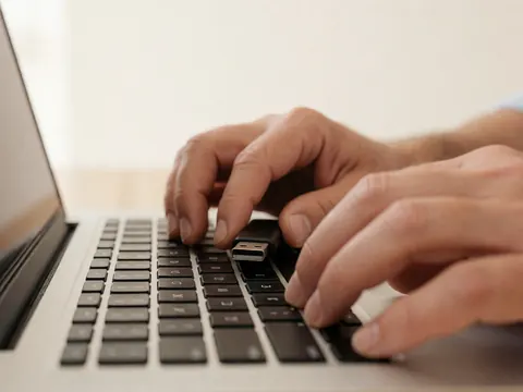 Close-up of hands typing on a laptop keyboard with a USB drive inserted, set against a warm white wall background.
