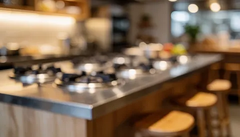 Blurred background of a modern kitchen island with stainless steel burners and wooden stools, featuring warm lighting and a cozy atmosphere suitable for text overlay.